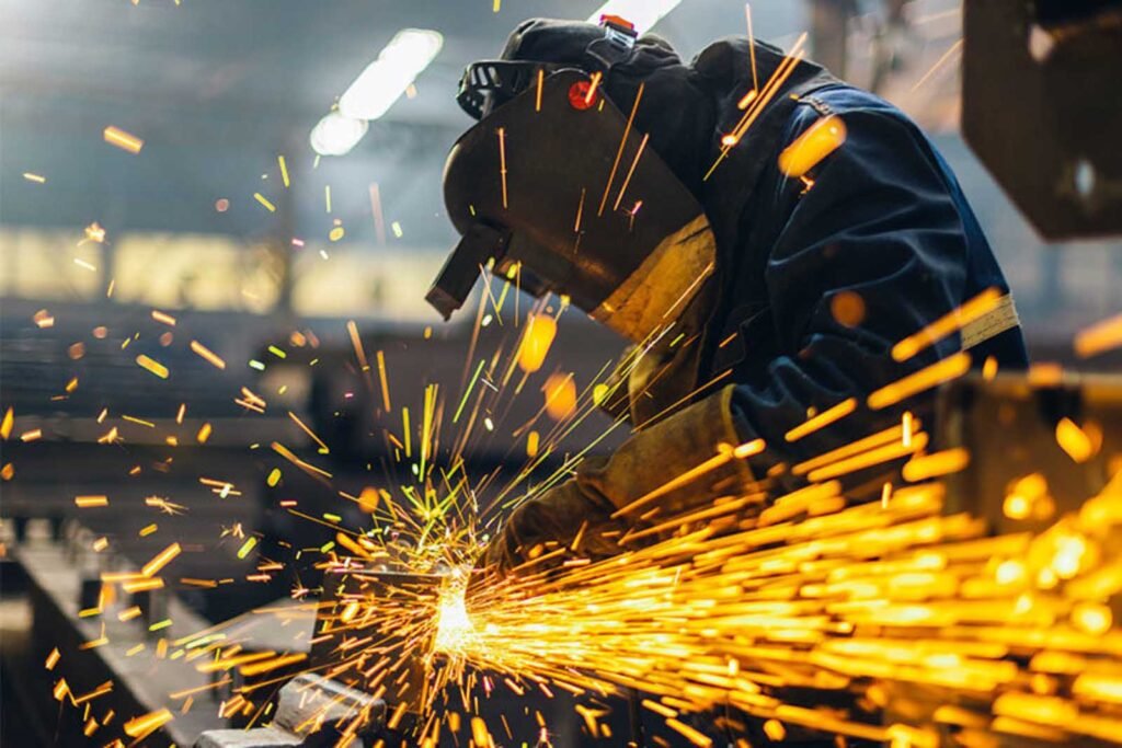 Industrial worker welding metal with protective gear and bright sparks flying during manufacturing process.